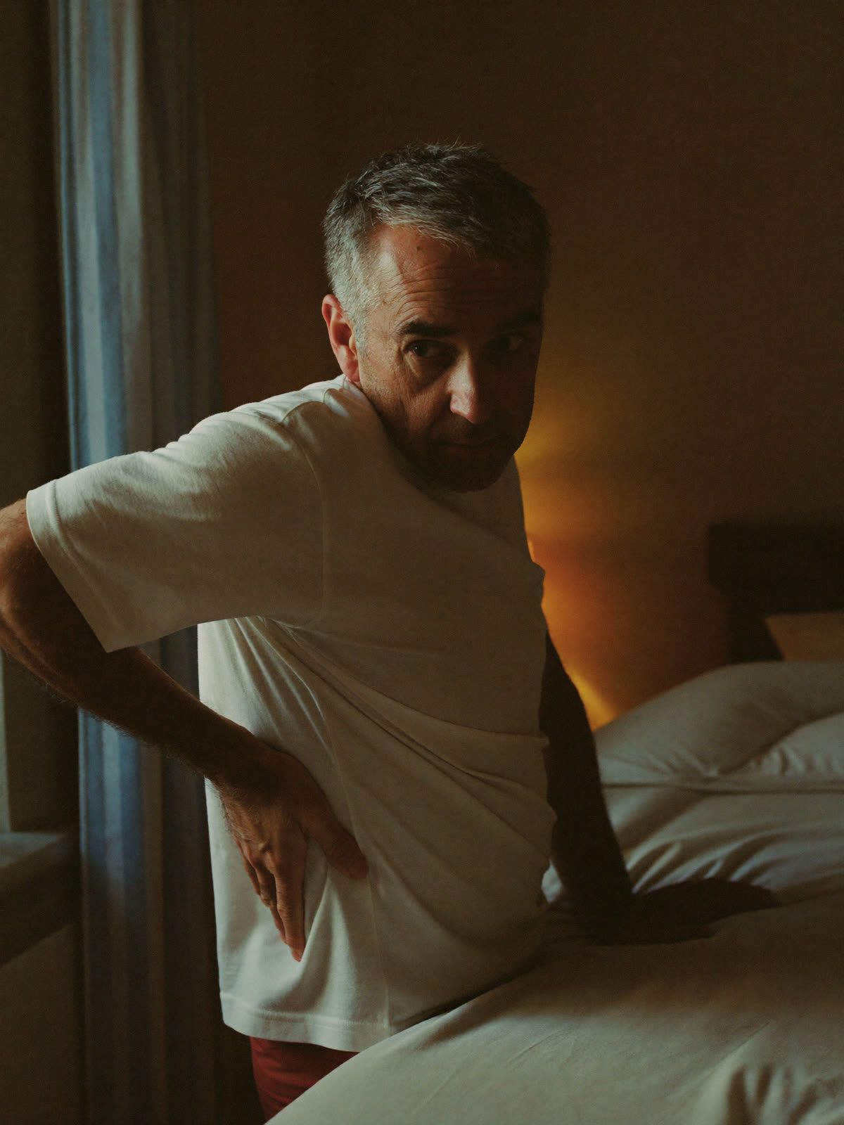A man in his fifties sitting on the edge of his bed at dawn, one hand pressed to his lower back