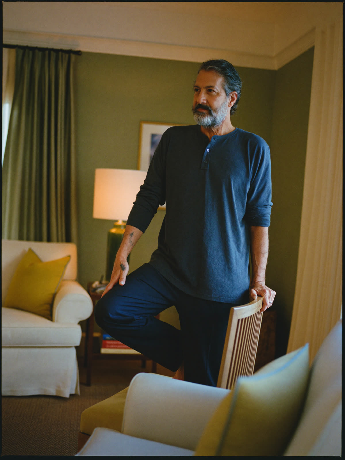 A man in his sixties doing a gentle standing movement at home, one hand on a chair for balance